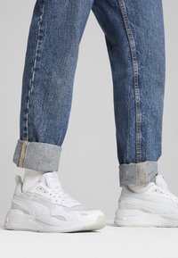 Personne portant des baskets blanches et un jean bleu retroussé, debout sur une surface grise claire unie.