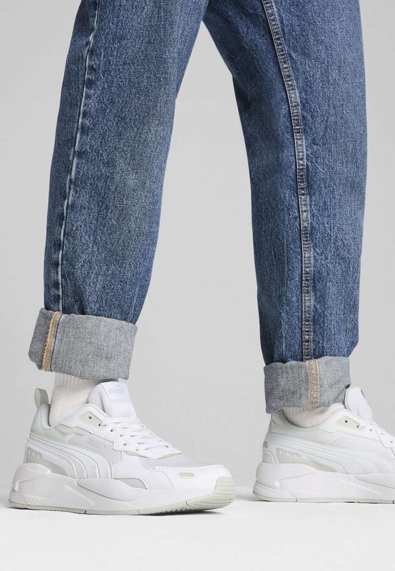 Personne portant des baskets blanches et un jean bleu retroussé, debout sur une surface grise claire unie.