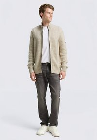 Hellbeige Strickjacke mit gerippter Struktur, Reißverschluss und langen Ärmeln, kombiniert mit einem weißen T-Shirt und dunklen Jeans, vor einem neutralen Hintergrund stehend.