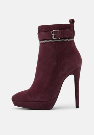 Bottine en daim bordeaux avec un bout pointu, un talon effilé, des détails en métal sur la fermeture éclair et un accent de boucle au niveau de la cheville. Texture lisse sur l'ensemble.