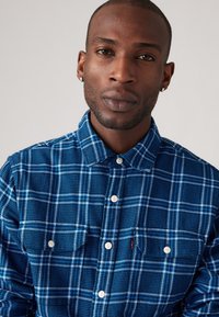 Chemise à carreaux bleus avec un col boutonné, dotée de deux poches poitrine et de boutons blancs. Fabriquée en tissu texturé avec des lignes contrastantes.