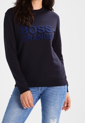 Femme portant un sweat-shirt bleu marine avec le texte « BOSS ORANGE » et un jean bleu clair déchiré, debout les mains détendues.