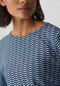 Eine Frau trägt ein Oberteil mit einem geometrischen Design in Blau, Weiß und Schwarz und einem runden Ausschnitt.