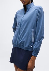 Γυναίκα που φοράει μπλε ζακέτα με φερμουάρ και ελαστικές μανσέτες, μαζί με μπλε σκούρο φούστα, στέκεται μπροστά σε λευκό απλό φόντο.