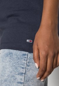 Navy cotton shirt with a small red, white, and blue logo stitched on the hem, paired with light blue denim jeans featuring a faded texture.