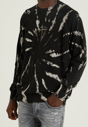 Czarna bluza tie-dye z długim rękawem z białym wzorem rozbłysku, mały biały napis na piersi, noszona z jasnoszarymi postrzępionymi dżinsami.