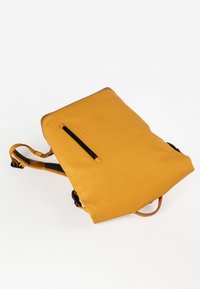 Zaino impermeabile giallo con una texture liscia, cinturini regolabili e una tasca con zip frontale. Presenta dettagli in pelle marrone sulla parte superiore.