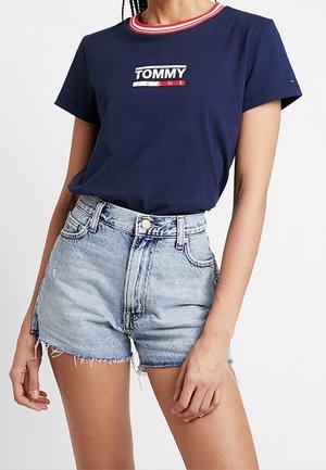 Žena nosící námořnicky modré tričko Tommy Jeans s červeným a bílým límečkem a světle modré vysoce sedící džínové šortky s roztřepenými lemy.