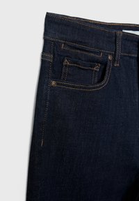 Gros plan sur un jean denim bleu foncé montrant la poche avant droite, la petite poche à pièces, les passants de ceinture, et les détails de couture orange.