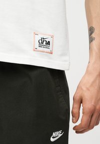 Λευκό πουκάμισο με το έμβλημα «Department of Nike Archives» πάνω από μαύρο παντελόνι Nike με λευκό λογότυπο, με ένα τατουάζ στο χέρι να ακουμπάει δίπλα.