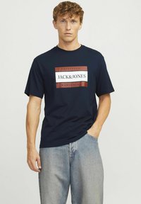 Marinblå bomulls t-shirt med en central logotypdesign i röd och vit. Korta ärmar och en avslappnad passform. Bärs med ljusblå jeans.