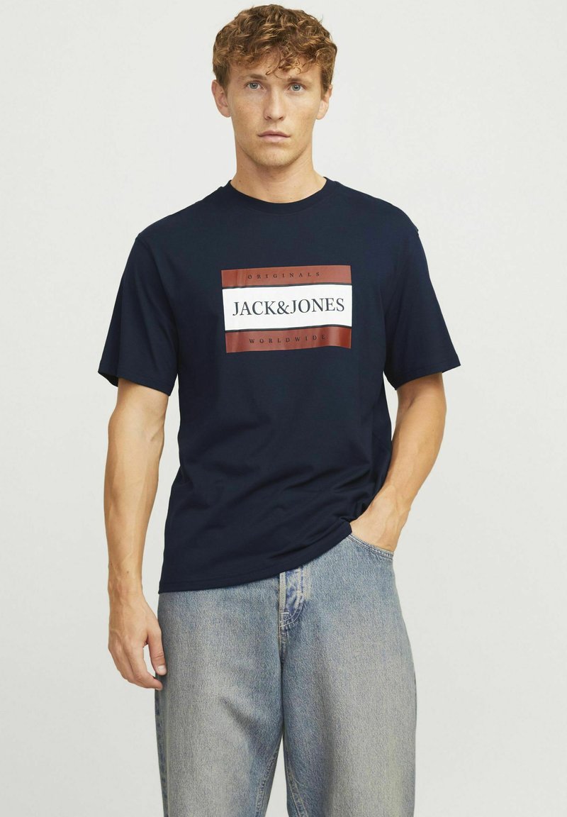 Marinblå bomulls t-shirt med en central logotypdesign i röd och vit. Korta ärmar och en avslappnad passform. Bärs med ljusblå jeans.