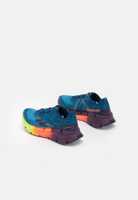 Zapatillas deportivas azules con parte superior de malla, que presentan acentos negros, una suela de degradado multicolor en amarillo, naranja y morado, y detalle de logo.