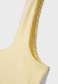 Detailní záběr na světle žlutý bezrukávový top z žebrované tkaniny, zobrazující ramenní řemínek a šev podpaží.