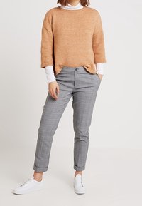 Hellbrauner gestrickter Pullover mit weiten Ärmeln, kombiniert mit grauen karrierten, schmal zulaufenden Hosen und weißen Sneakern. Gemütliches und lässiges Outfit.