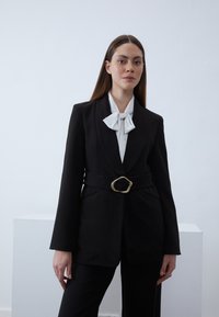 Vrouw draagt een zwarte blazer met riem over een witte blouse met een grote strik, staande tegen een eenvoudige lichte achtergrond.