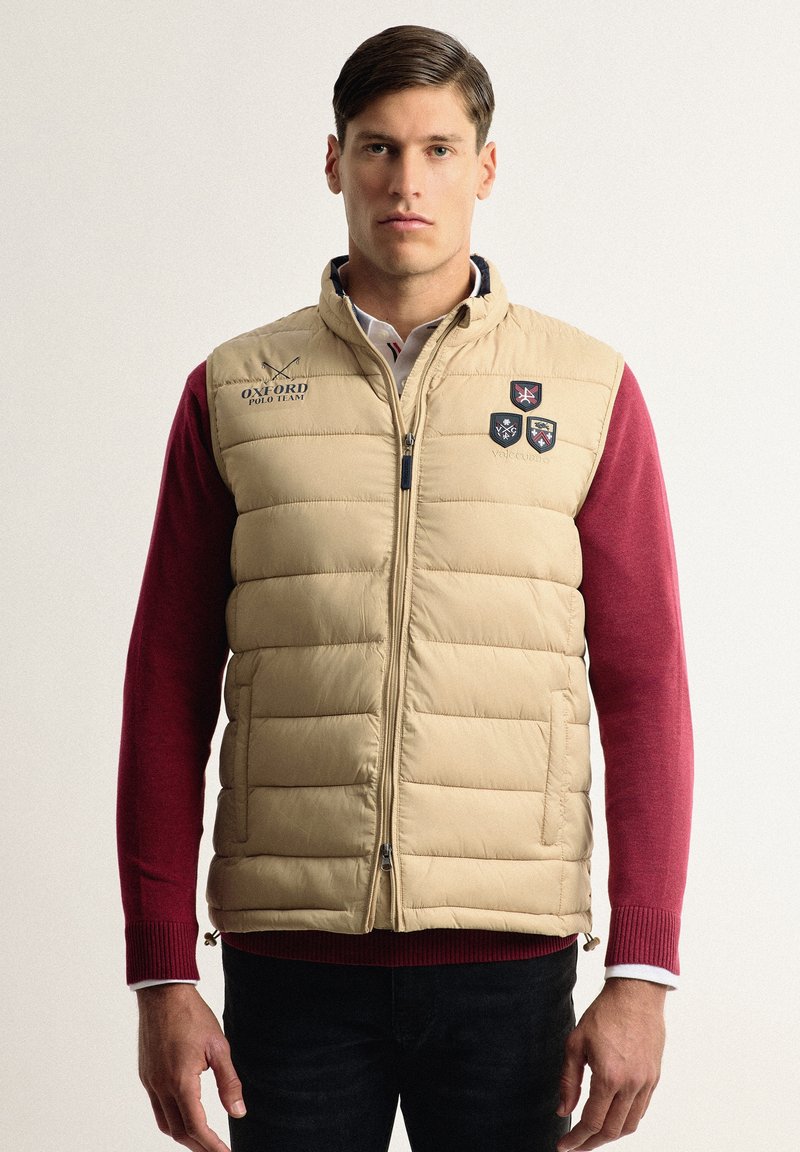 Chaleco de plumas beige con costuras horizontales, cremallera frontal y detalles de logotipo. Llevarlo sobre una camiseta de manga larga roja, que presenta un cuello.