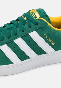 Green suede sneaker with white leather three stripes, yellow accents, and perforations for breathability. Features a cushioned sole.