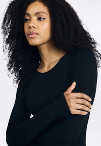 Mulher jovem com cabelo longo e encaracolado, vestindo uma camisa de manga longa preta, olhando para o lado com o braço esquerdo cruzado sobre o corpo.
