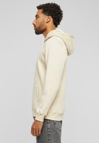 Beige hettegenser med kengurulomme og ribbestrikkede mansjetter. Stoffet virker mykt, og designet er enkelt og minimalistisk.