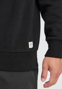 Sudadera negra con puños y dobladillo acanalados. Presenta una pequeña etiqueta de tela en el costado. El material parece suave y texturizado.