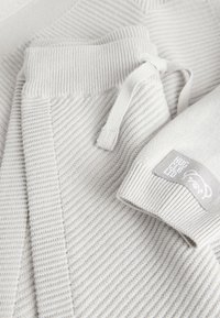 Suéter de punto gris claro con textura acanalada, cordón ajustable en la cintura y parche de marca en la manga.