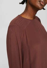 Blusa color ruggine con maniche lunghe, caratterizzata da un colletto plissettato testurizzato e un tessuto morbido e fluido. Collo rotondo, texture liscia.