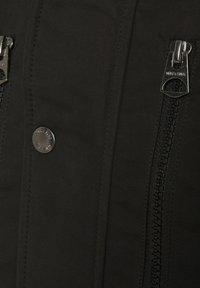 Svart tygjacka med en slät yta, dubbla blixtlåsfickor, en metallknapp och kontrasterande blixtlåsdetaljer.