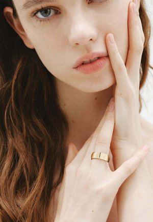 Joven con ojos azules y cabello castaño largo tocándose suavemente el rostro, llevando un anillo de oro con pequeñas gemas en el dedo.