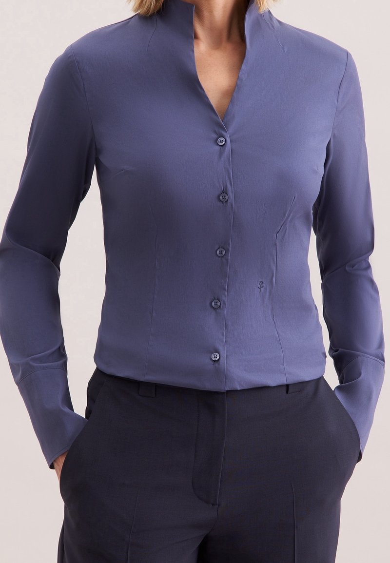 Chemise bleue ajustée à manches longues avec boutons, portée avec un pantalon sombre sur mesure, portée par une personne les mains dans les poches.