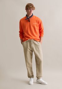 Pomarańczowa bluza z prążkowanymi mankietami, beżowe spodnie dresowe ze ściągaczami i kieszeniami po bokach oraz białe trampki. Akcesorium na szyję w postaci bandany.