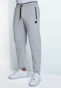 Graue Jogginghosen mit schmal zulaufendem Design, ausgestattet mit einer vorderen Reißverschlusstasche, kontrastierendem schwarzen Kordelzug und dehnbaren Bündchen.