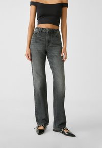 Donna che indossa jeans neri sbiaditi a vita alta, un crop top nero off-shoulder e ballerine nere a punta con cinturino, in piedi su uno sfondo semplice.