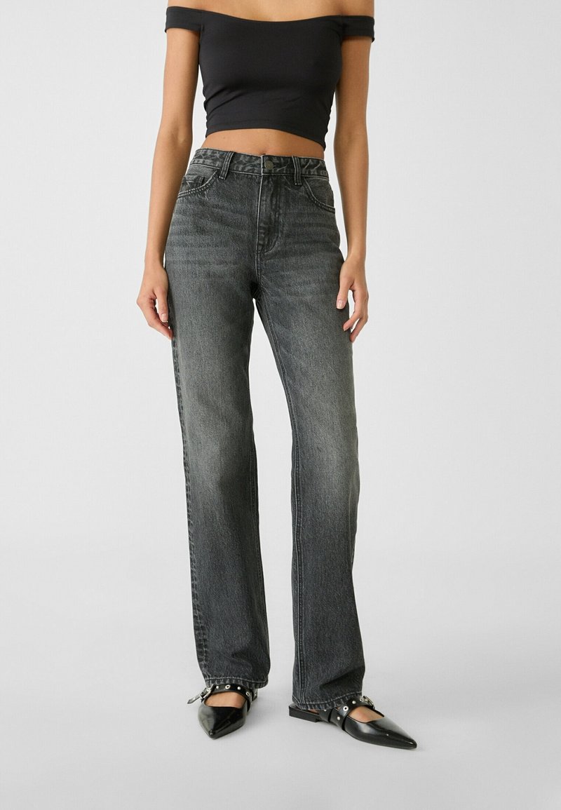 Donna che indossa jeans neri sbiaditi a vita alta, un crop top nero off-shoulder e ballerine nere a punta con cinturino, in piedi su uno sfondo semplice.