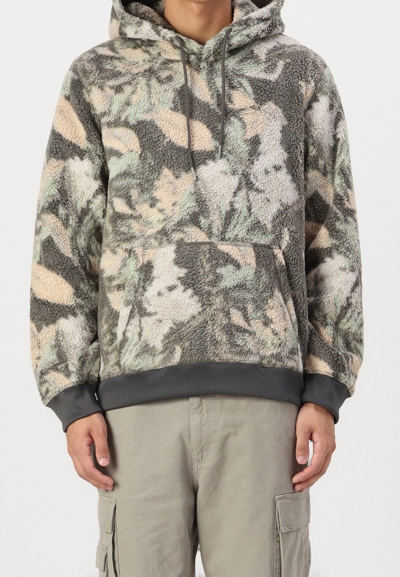 Personne portant un sweat à capuche en polaire à motif camouflage vert, gris et beige avec poche kangourou et poignets et ourlet côtelés gris.
