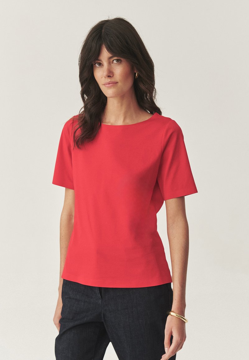 Tricou roșu cu mâneci scurte și guler rotund, fabricat dintr-un material neted, asortat cu pantaloni din denim închis. Design simplu, fără modele sau accente.