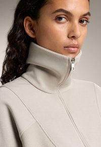 Béžová mikina s vysokým golierom a zipsom. Obsahuje ribbované textúry na pleciach a hladký povrch. Jednoduchý dizajn s minimálnymi akcentmi.