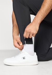 Zwarte sportieve broek met een zijritsdetail, gedragen met witte sneakers en geribbelde witte sokken, wat een sportieve, functionele uitstraling benadrukt.