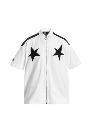 Joven con pantalones cortos blancos y camisa de manga corta con grandes estrellas negras, camiseta interior negra, calcetines blancos y zapatos beige con textura.