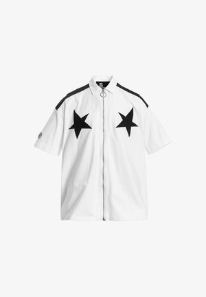 Joven con pantalones cortos blancos y camisa de manga corta con grandes estrellas negras, camiseta interior negra, calcetines blancos y zapatos beige con textura.