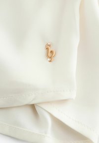 Blouse couleur crème avec une texture lisse, ornée d'une petite étiquette en métal doré avec un design en cursive cousu sur le tissu.