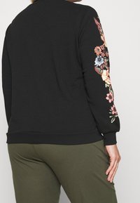 Czarny sweter z kolorową haftowaną florystyką na lewym rękawie, z ściągaczami na mankietach i u dołu. W zestawie z oliwkowymi spodniami.