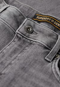 La tela de mezclilla gris presenta un cierre de botón, un herraje de metal y una etiqueta con "Superdry Denim Goods" en naranja y negro.