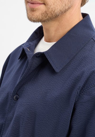 Homme portant une chemise à boutons texturée bleu marine par-dessus un t-shirt blanc à col rond, montrant le cou, le menton et une barbe partielle.