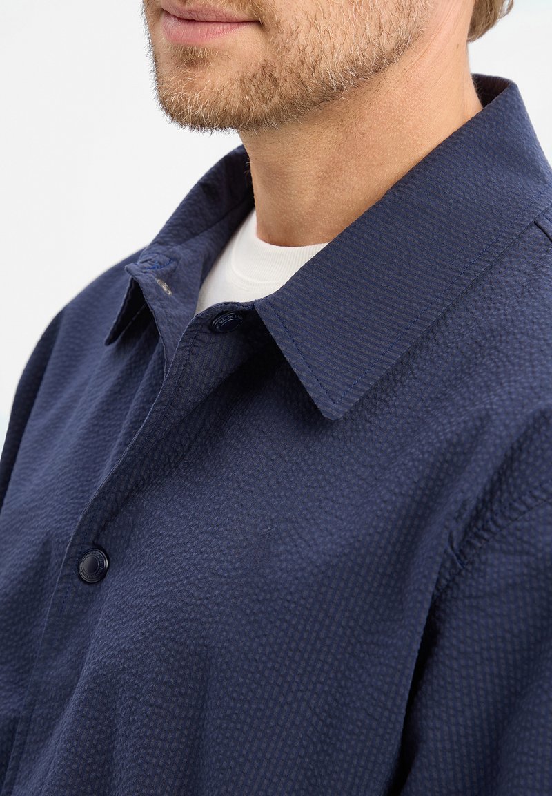 Homme portant une chemise à boutons texturée bleu marine par-dessus un t-shirt blanc à col rond, montrant le cou, le menton et une barbe partielle.