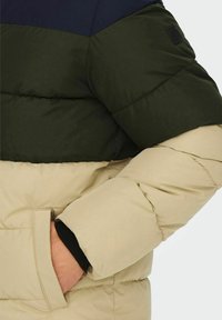 Bunda s kapucňou so štruktúrovanou látkou, ktorá má tmavomodrú hornú časť, tmavozelené stredné pásmo a béžovú spodnú časť. Obsahuje bočnú kapsu a elastické manžety.