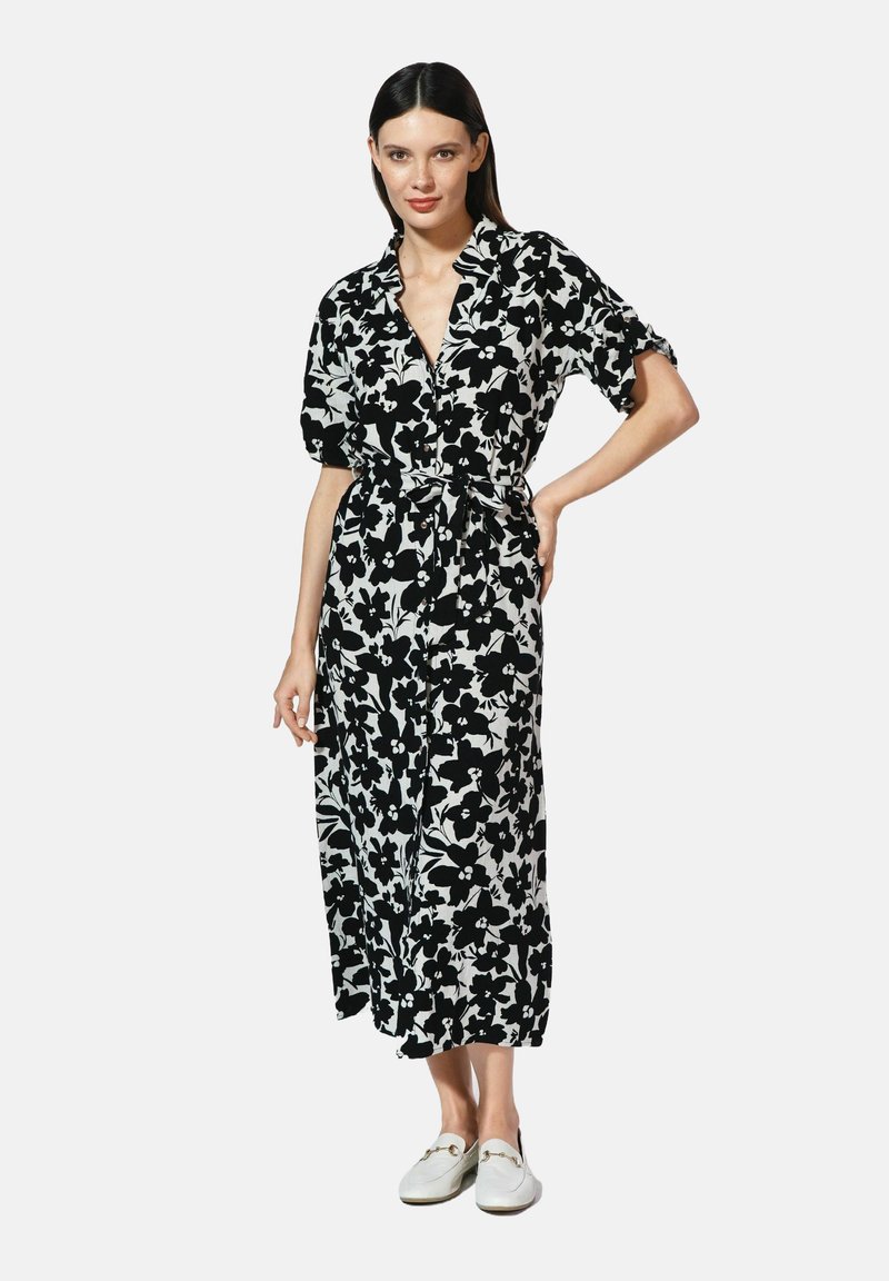 Czarna i biała kwiatowa maxi sukienka z krótkimi rękawami, zapięciem na guziki z przodu i paskiem w talii. Modelka nosi białe slip-on buty.