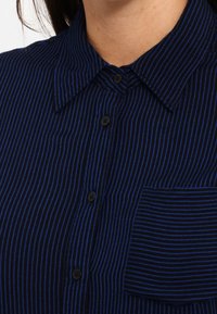 Koszula zapinana na guziki w ciemnoniebieskim kolorze z cienkimi pionowymi paskami. Posiada kołnierzyk, czarne guziki oraz kieszeń w dopasowanym wzorze pasków.