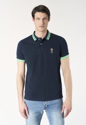 Uomo che indossa una polo blu navy con colletto e polsini a righe verdi, in piedi davanti a uno sfondo semplice con jeans azzurri.