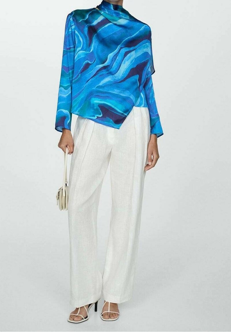 Personne portant un haut à manches longues bleu à motifs, un pantalon blanc à jambes larges, des sandales blanches à talons, et tenant un petit sac à main beige.